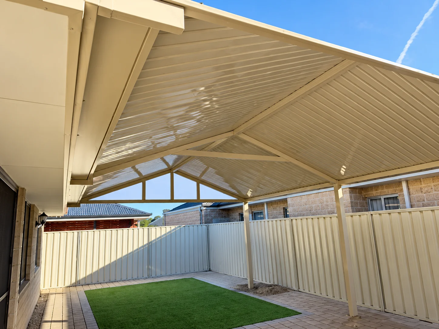 Gable patio roof installation over a paved backyard with artificial turf.